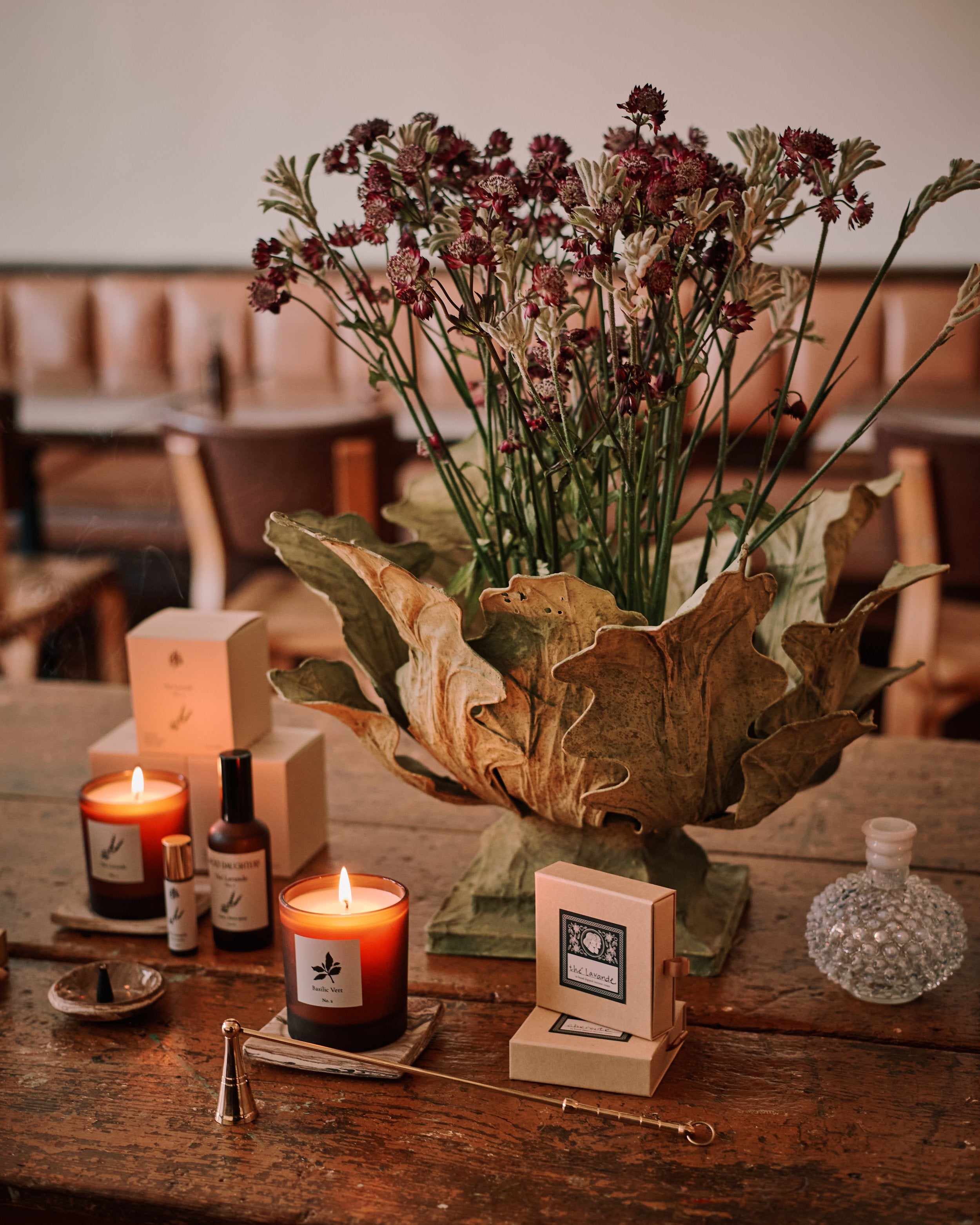A collection of candles and incense boxes with a flower arrangement in a leafy container on a wood table.
