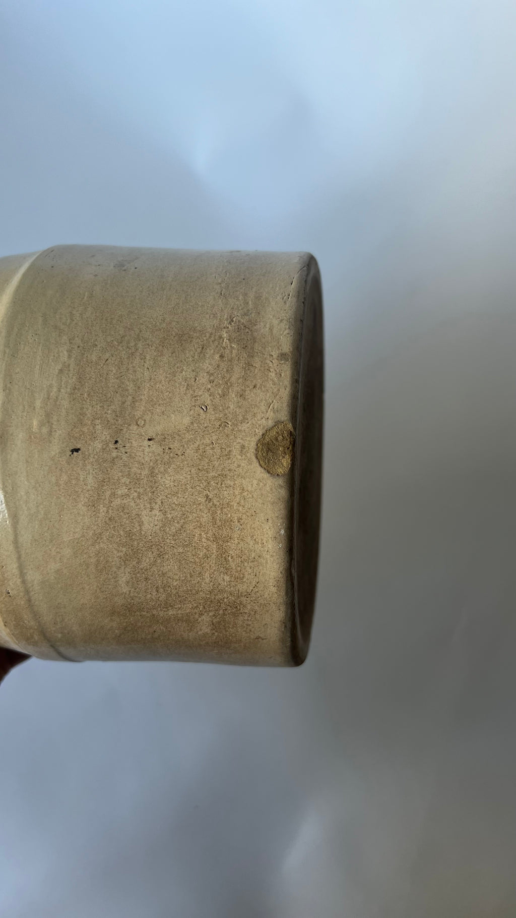 A close-up of a cylindrical ceramic jug  with a rustic, weathered finish, held by a hand against a plain light gray background.