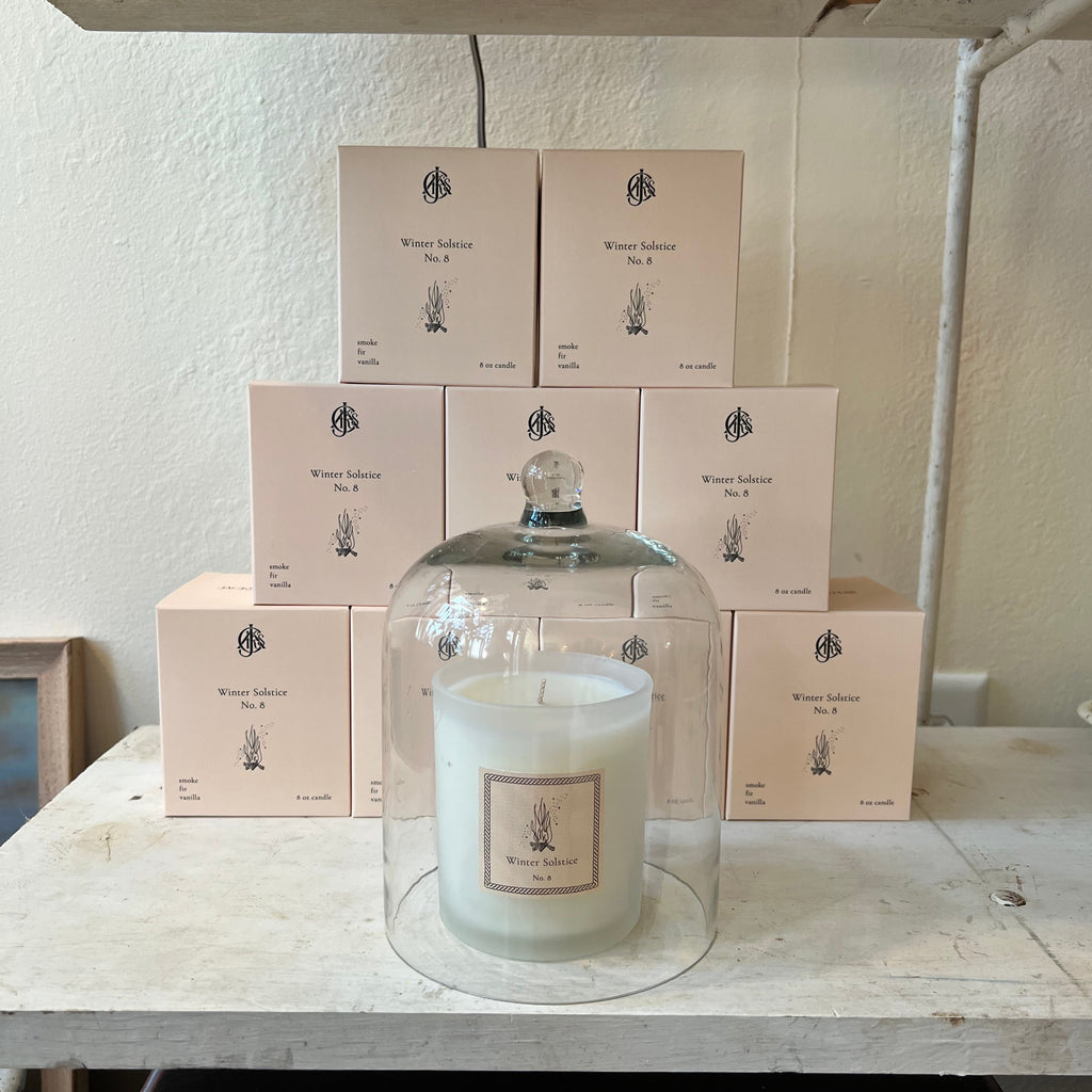 Candle in a glass cloche with a stack of candle boxes on a wooden shelf.