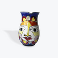 A hand-painted ceramic pitcher with a stylized face design, featuring blue, yellow, red, and white colors, set against a plain light gray background.