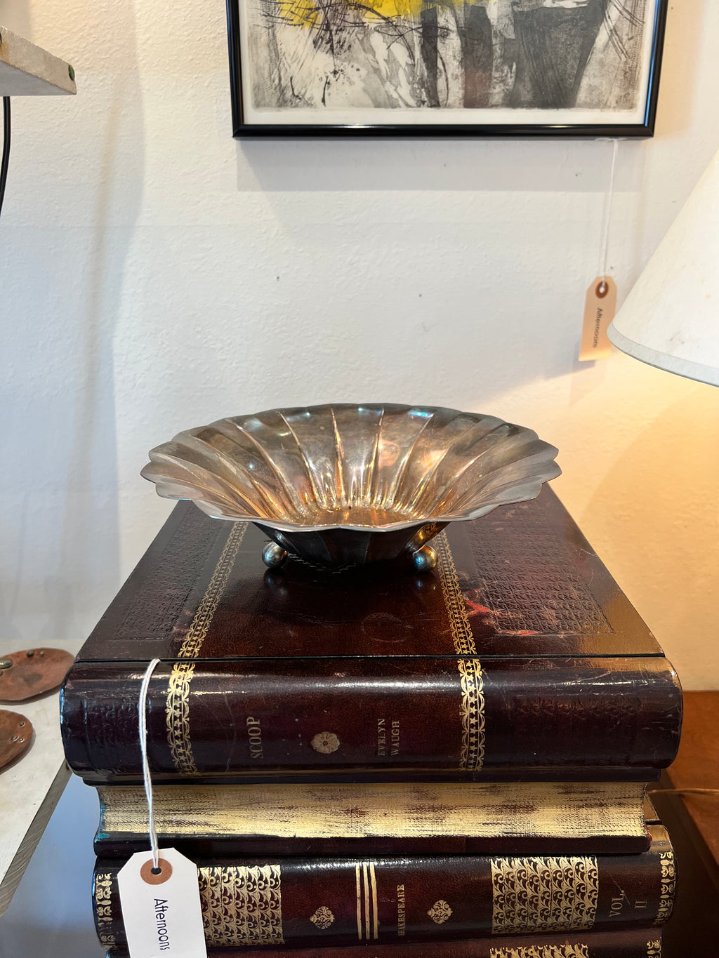 A stack of vintage books with a decorative silver footed bowl on top and framed picture in the background.
