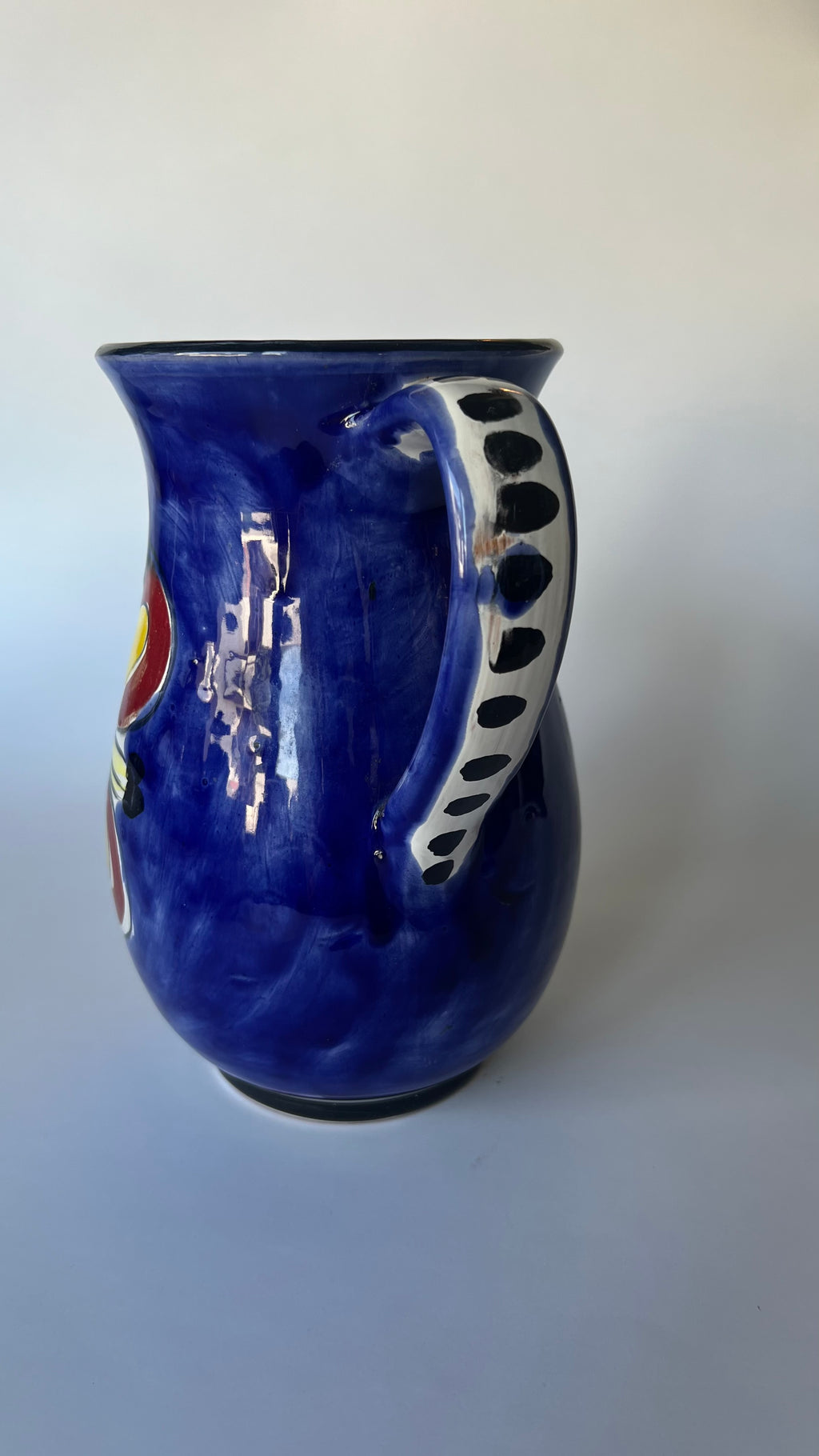 A glossy cobalt blue ceramic pitcher with a white handle decorated with black polka dots and a small multicolor emblem on the side, set against a plain light gray background.