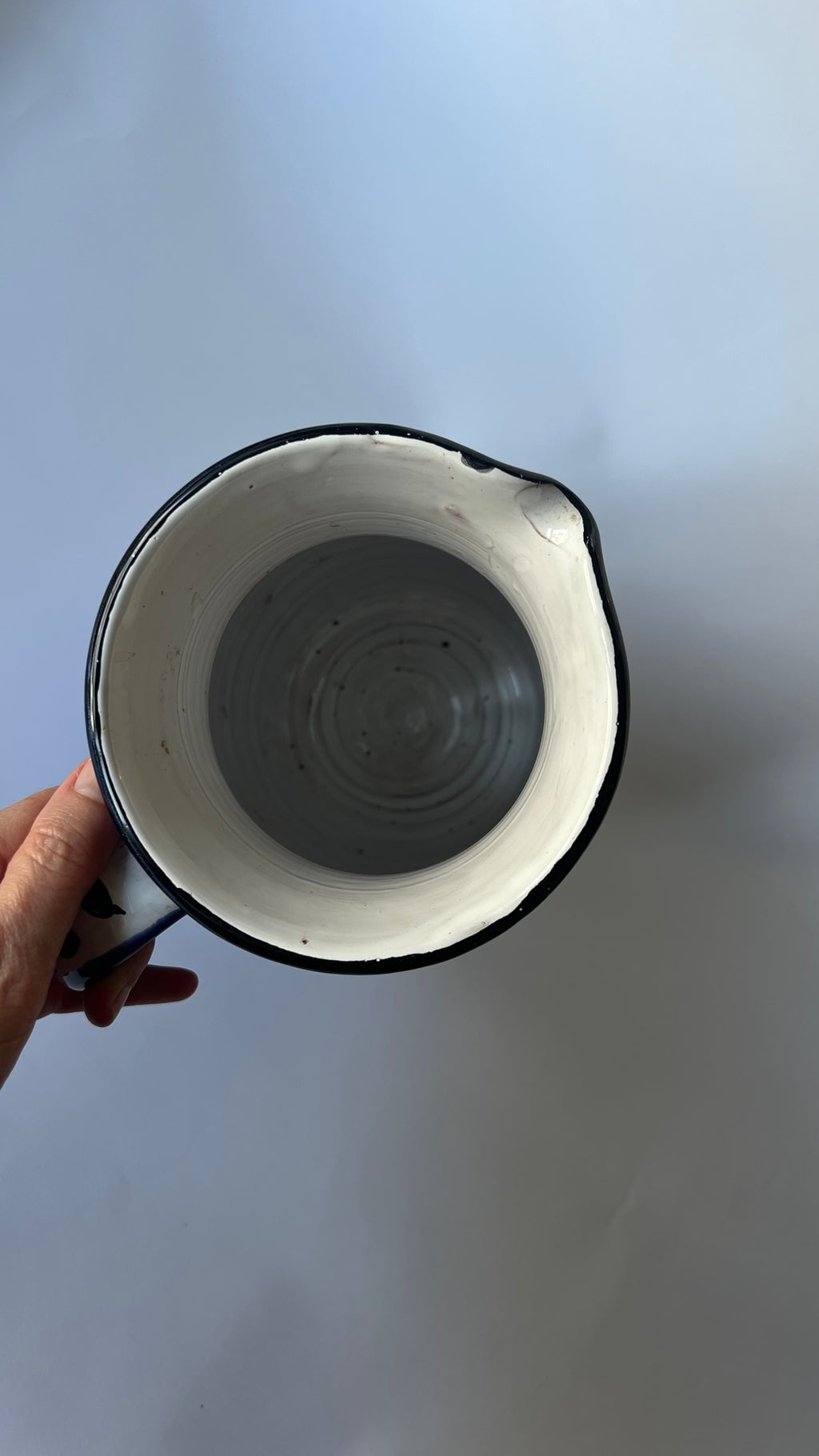 A hand holding a handmade ceramic pitcher with a white interior, black rim, and a gray glazed center, set against a plain light gray background.