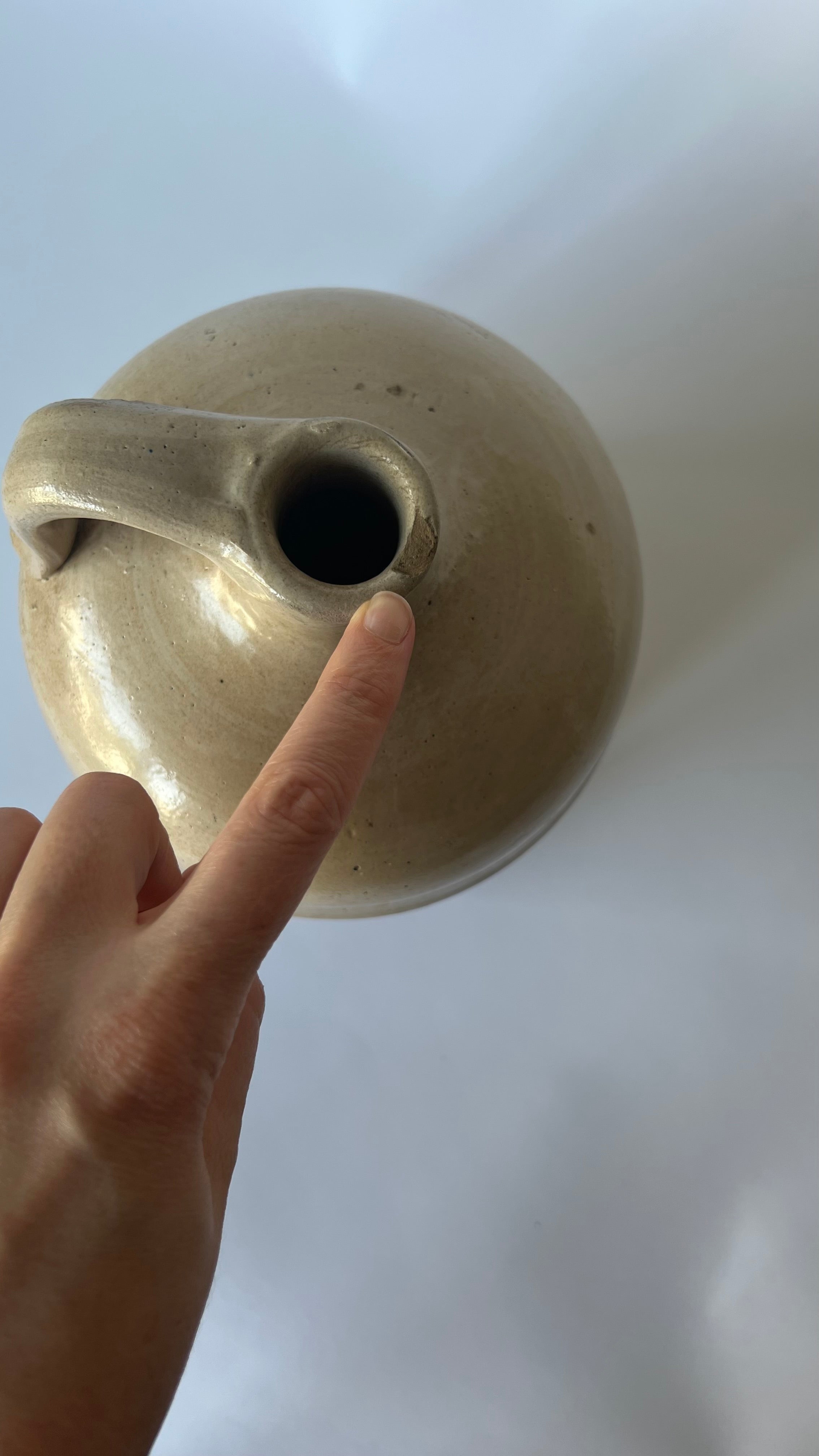 A hand pointing at the opening of a round, beige ceramic jug with a handle, set against a plain light background.