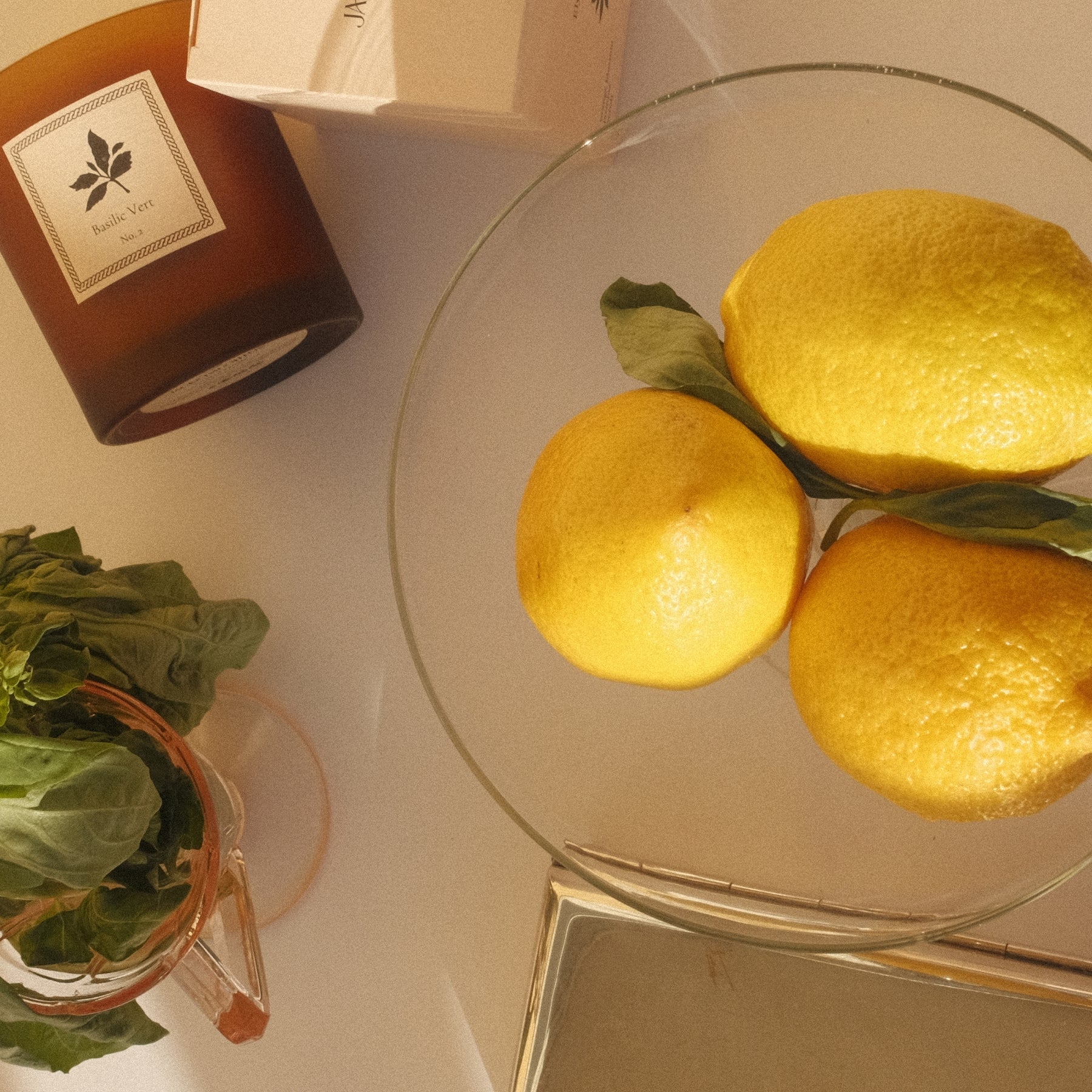 Three lemons on a glass plate with a basilic vert candle and a container of herbs in the background.
