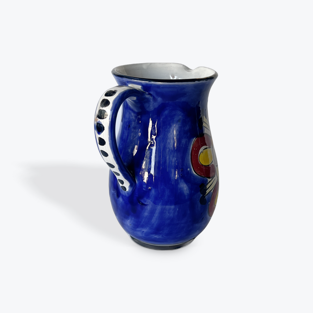 A cobalt blue ceramic pitcher with a white handle featuring black polka dots, and a colorful red, yellow, and white design on the side, set against a plain light gray background.