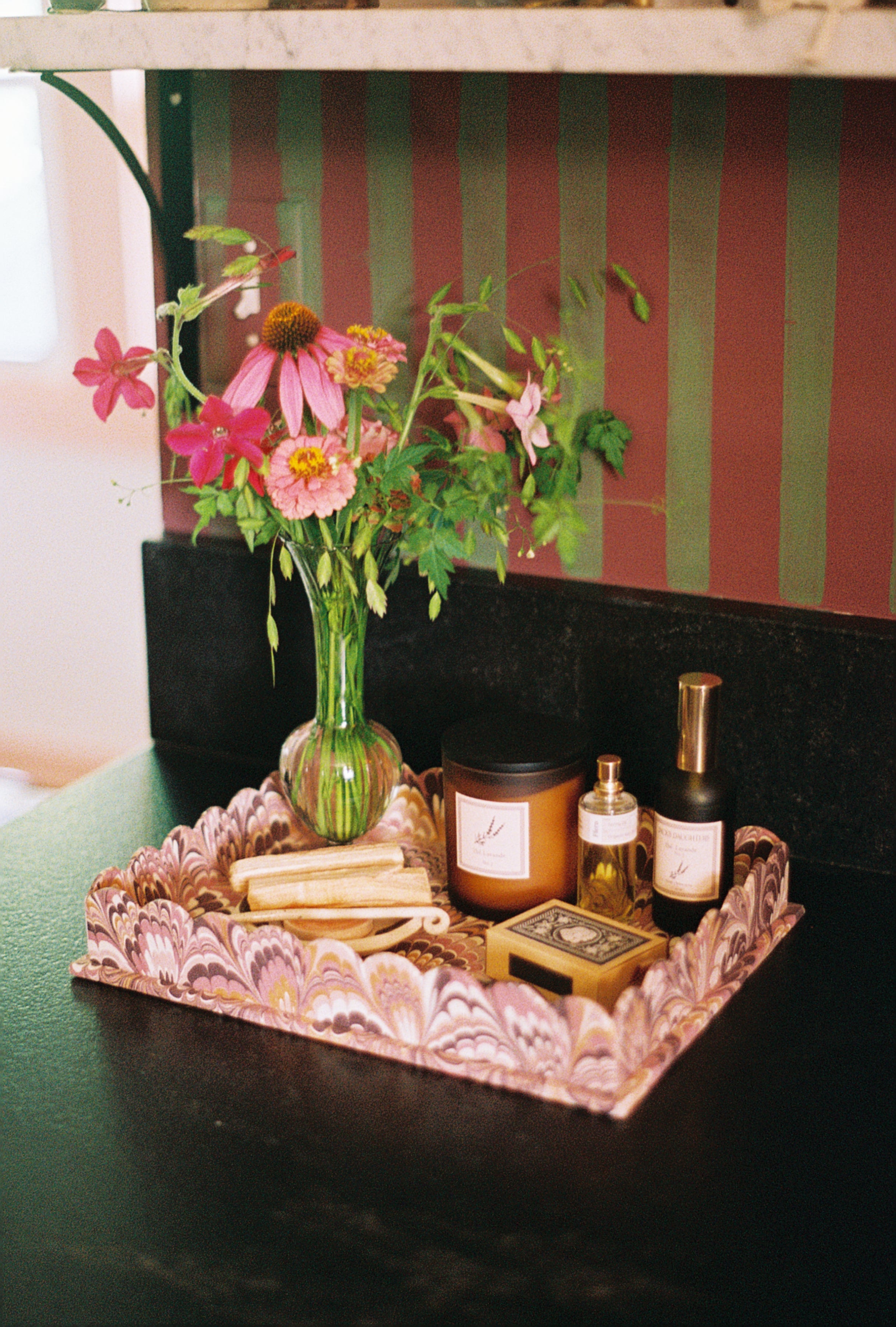 A decorative pink and white patterned tray holds a glass vase with pink and orange flowers, a candle jar, a small bottle, and a matchbox on a dark tabletop. A striped wall in muted red and green is visible in the background.
