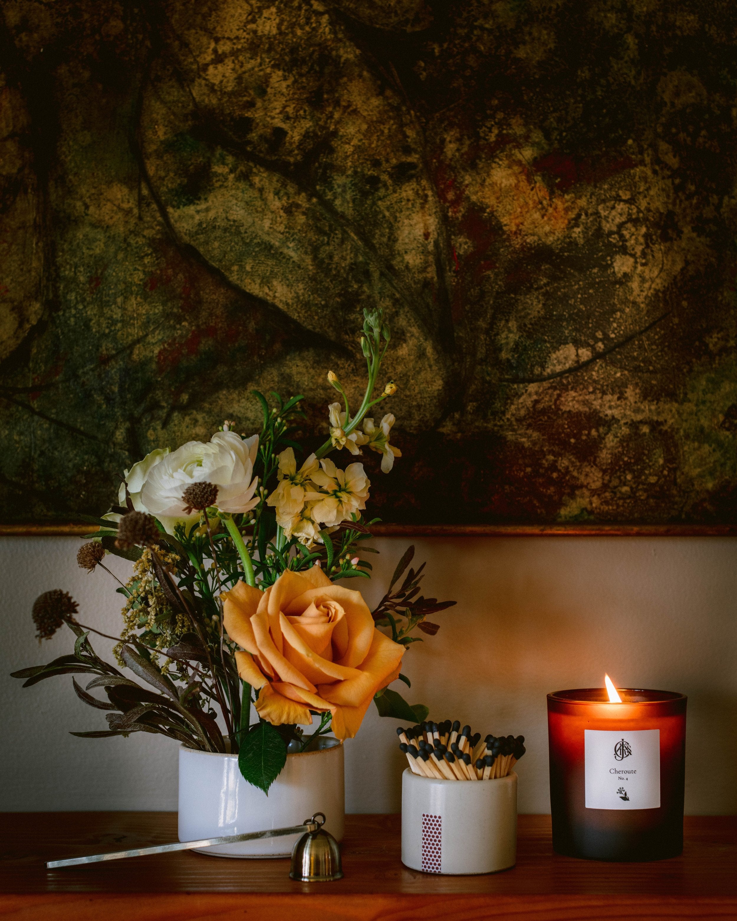 Floral arrangement with a lit candle and matches on a wooden surface against a textured wall.