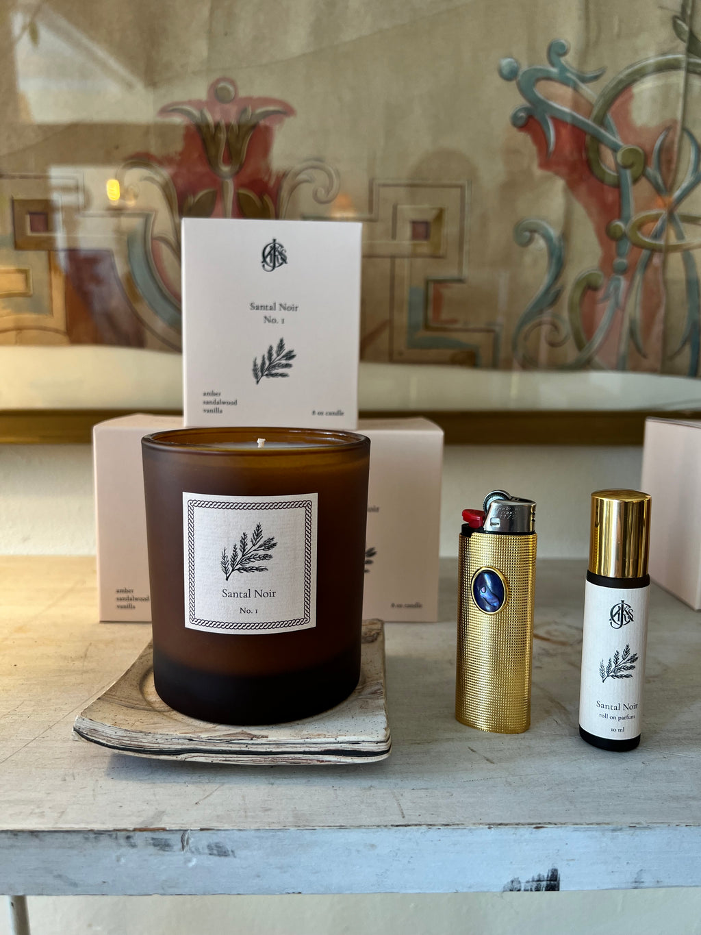 A candle, lighter case and roll on perfume on a wooden surface with a patterned background