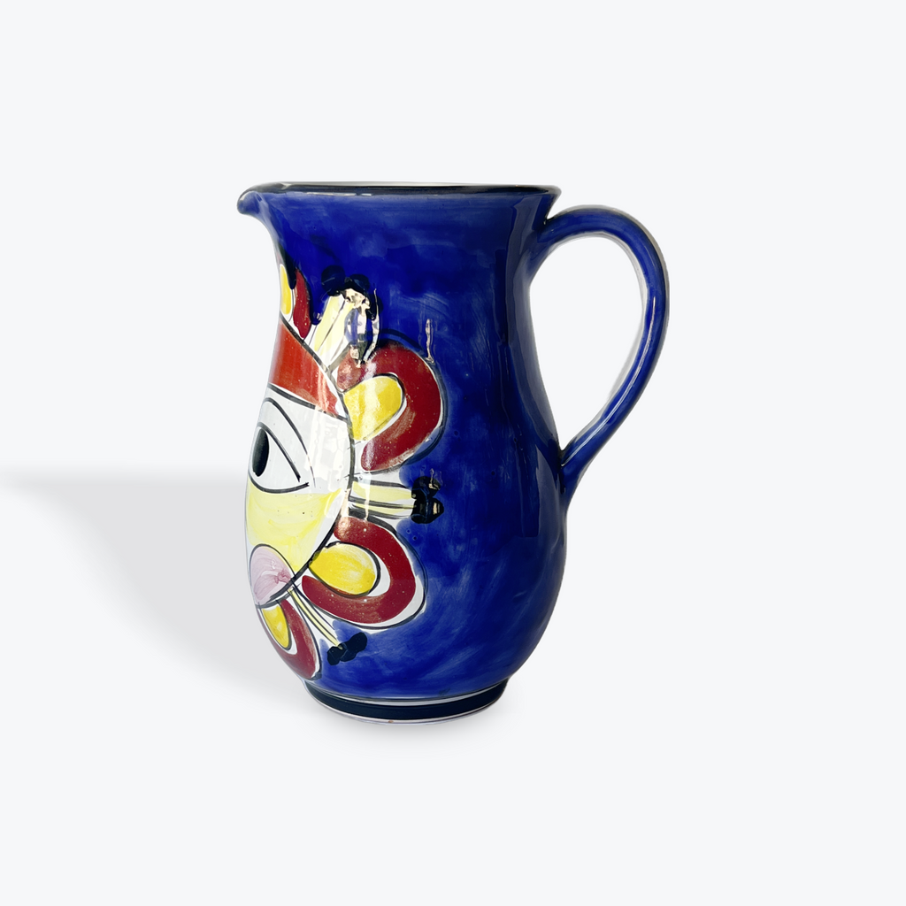 A cobalt blue ceramic pitcher with a colorful hand-painted face design on the side, set against a plain light gray background.
