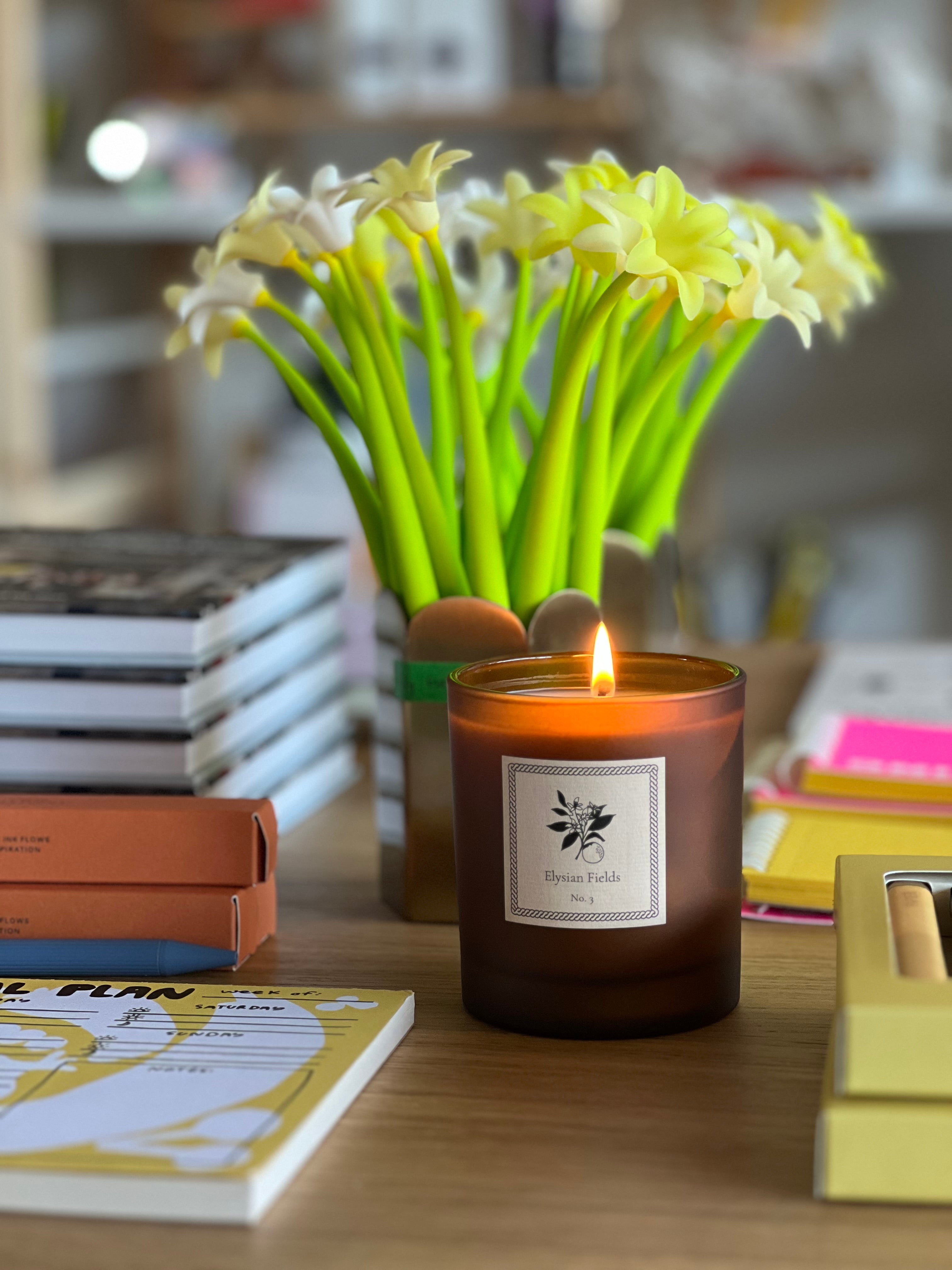 Candle with a label on a table next to a vase of flowers and books