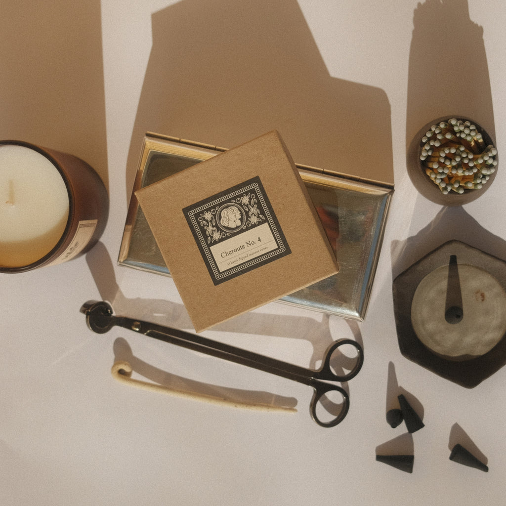 Collection of decorative items including a candle, incense box, wick trimmers, matches and incense dish on a white surface with shadows.  
