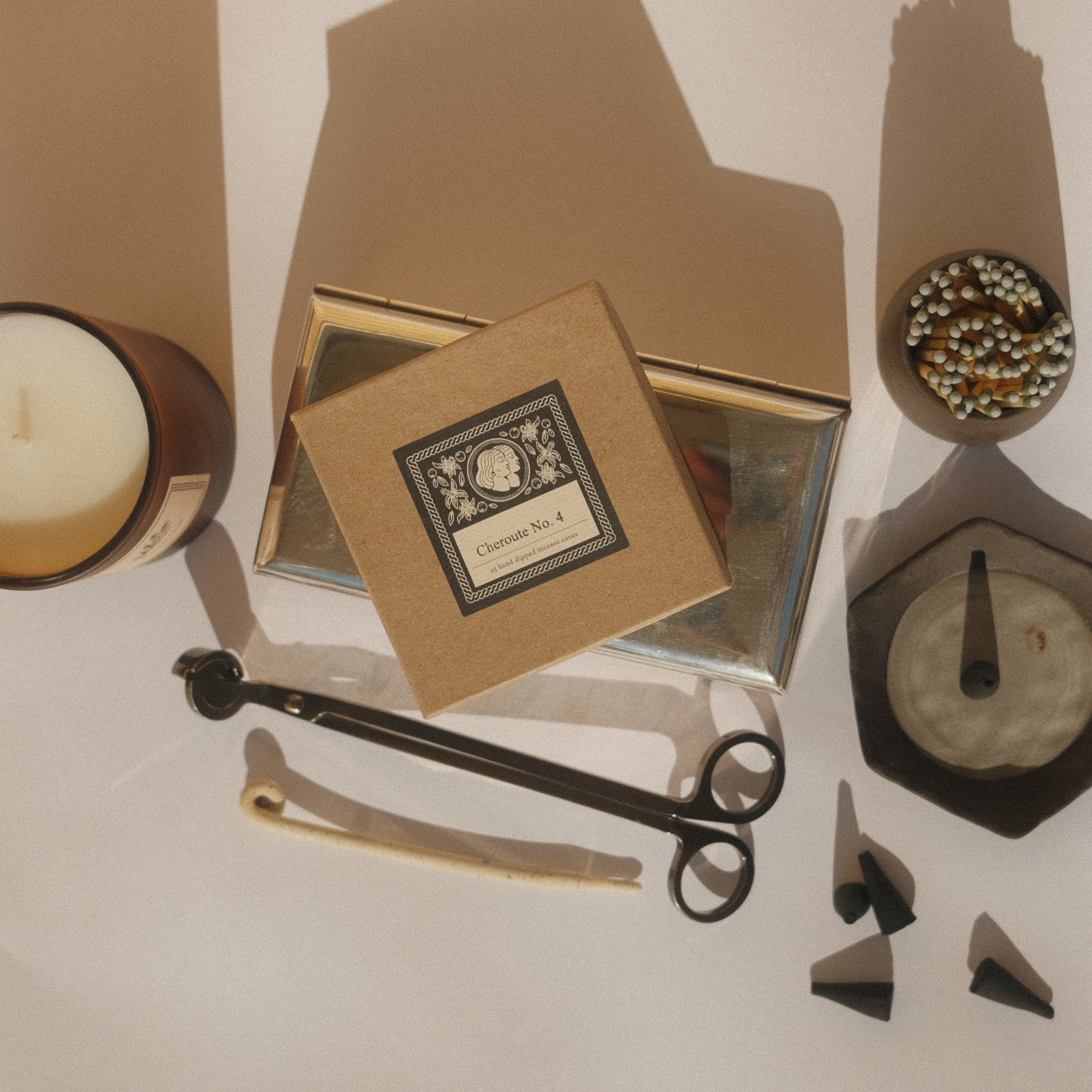 Collection of decorative items including a candle, incense box, wick trimmers, matches and incense dish on a white surface with shadows.  