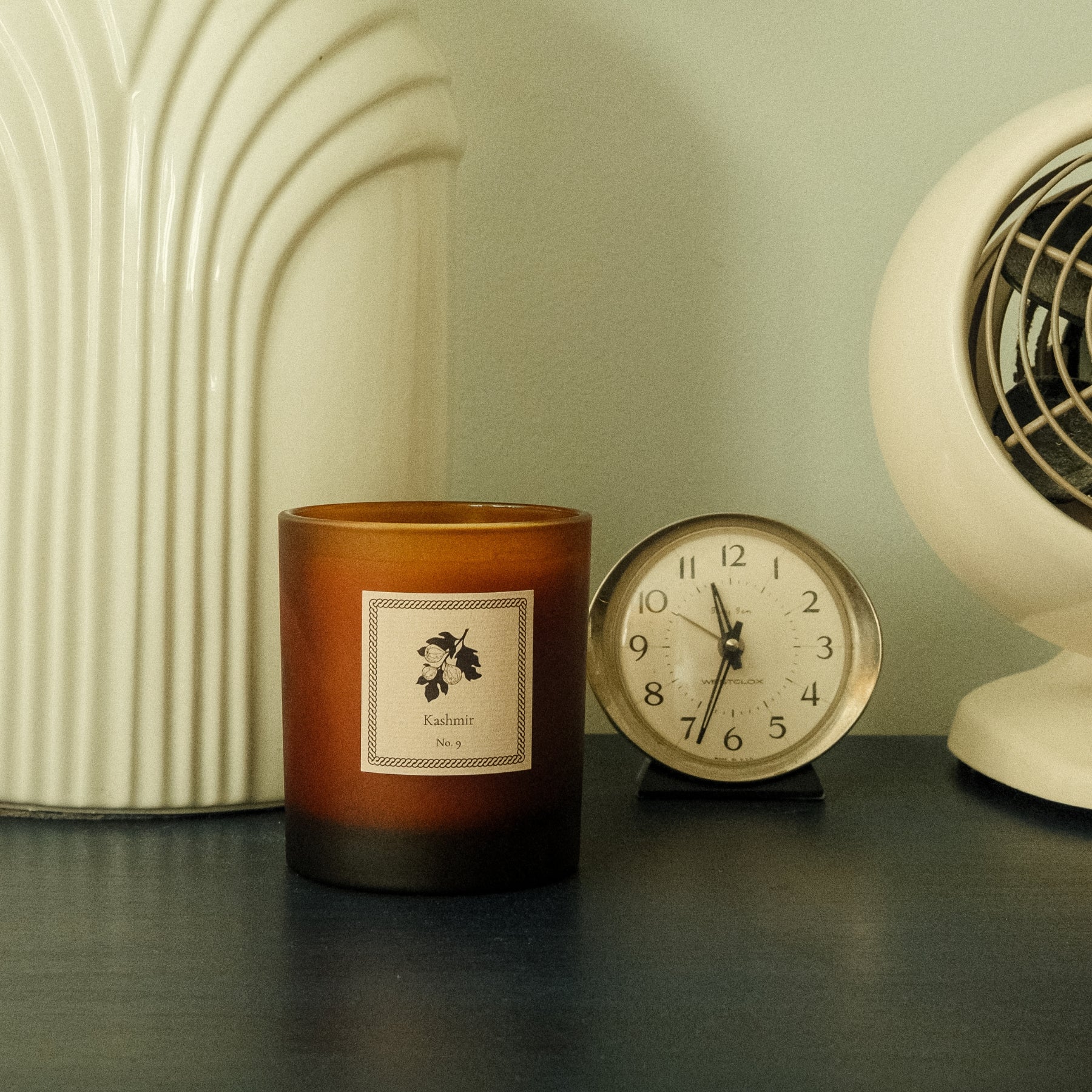 Candle with a label, clock, and vintage-style fan on a surface.