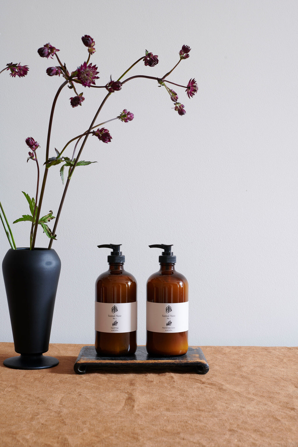 Two amber glass bottles with black pump dispensers on a wooden surface, accompanied by a black vase with purple flowers.
