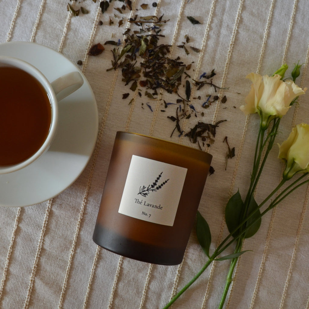 Candle labeled 'The Lavande' with a cup of tea and flowers on a textured surface