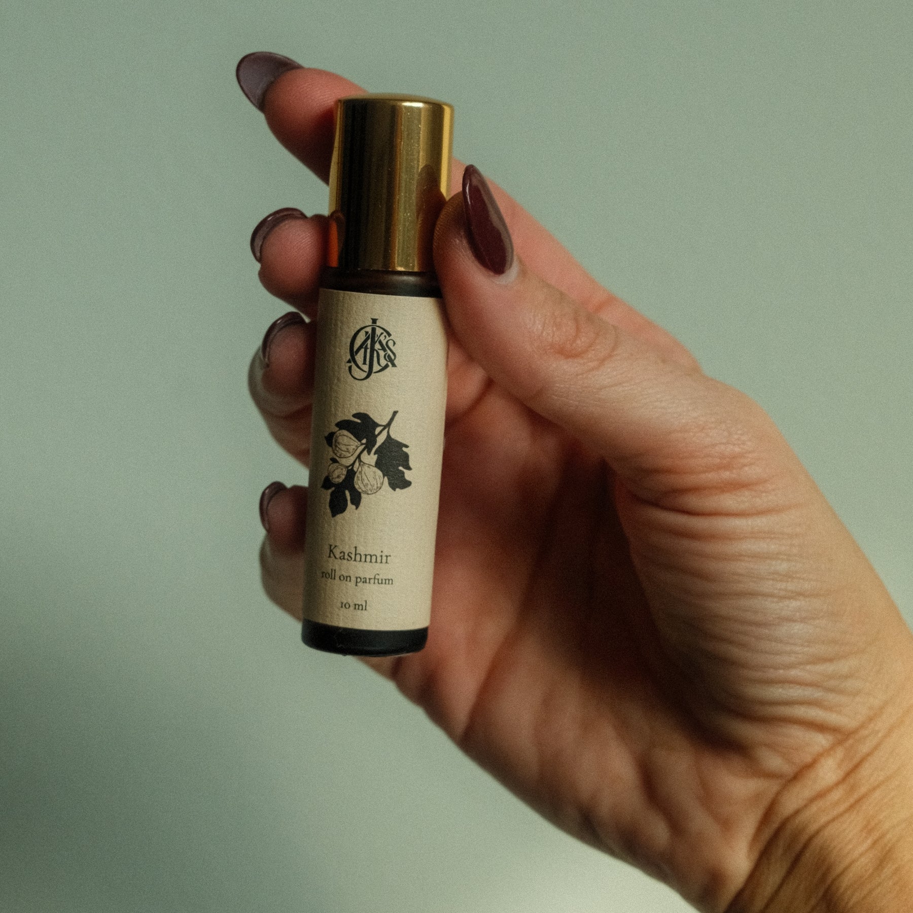 Hand holding a roll on perfume bottle labeled 'Kashmir' with a gold cap against a light green background