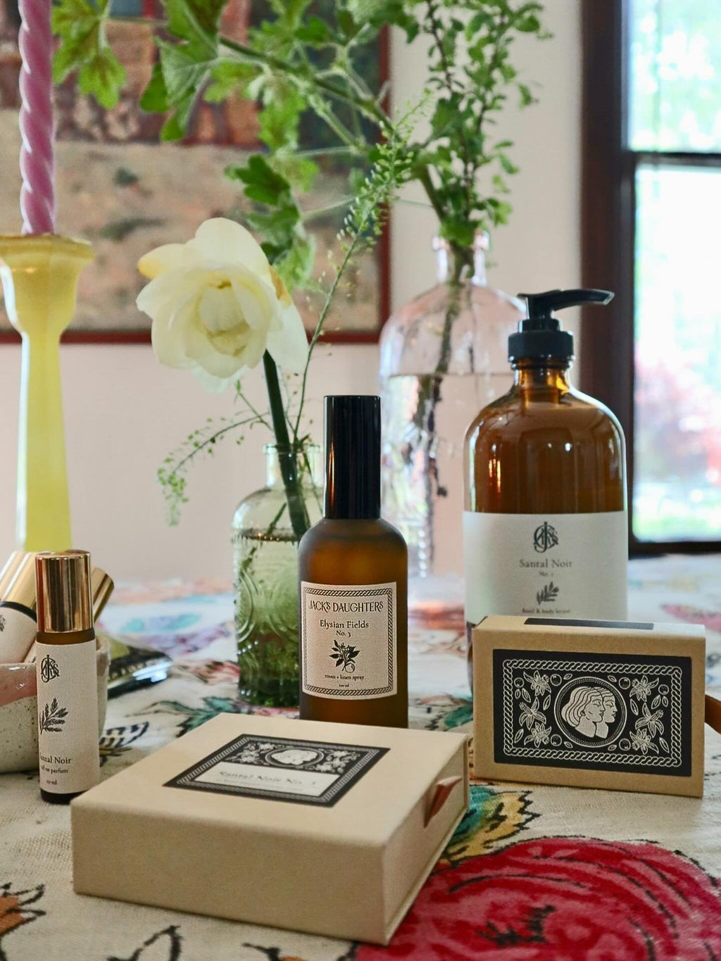 Collection of products including bottles and a matchbox on a table with a floral arrangement in the background.