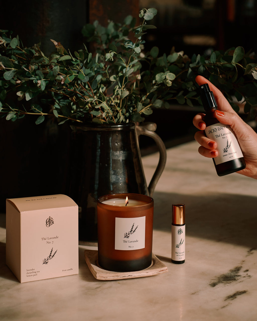 Candle, packaging, and bottles on a marble surface with greenery in the background