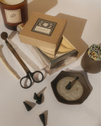 Candle tools and materials including a candle, wick trimmer, and a ceramic incense  holder on a neutral background.