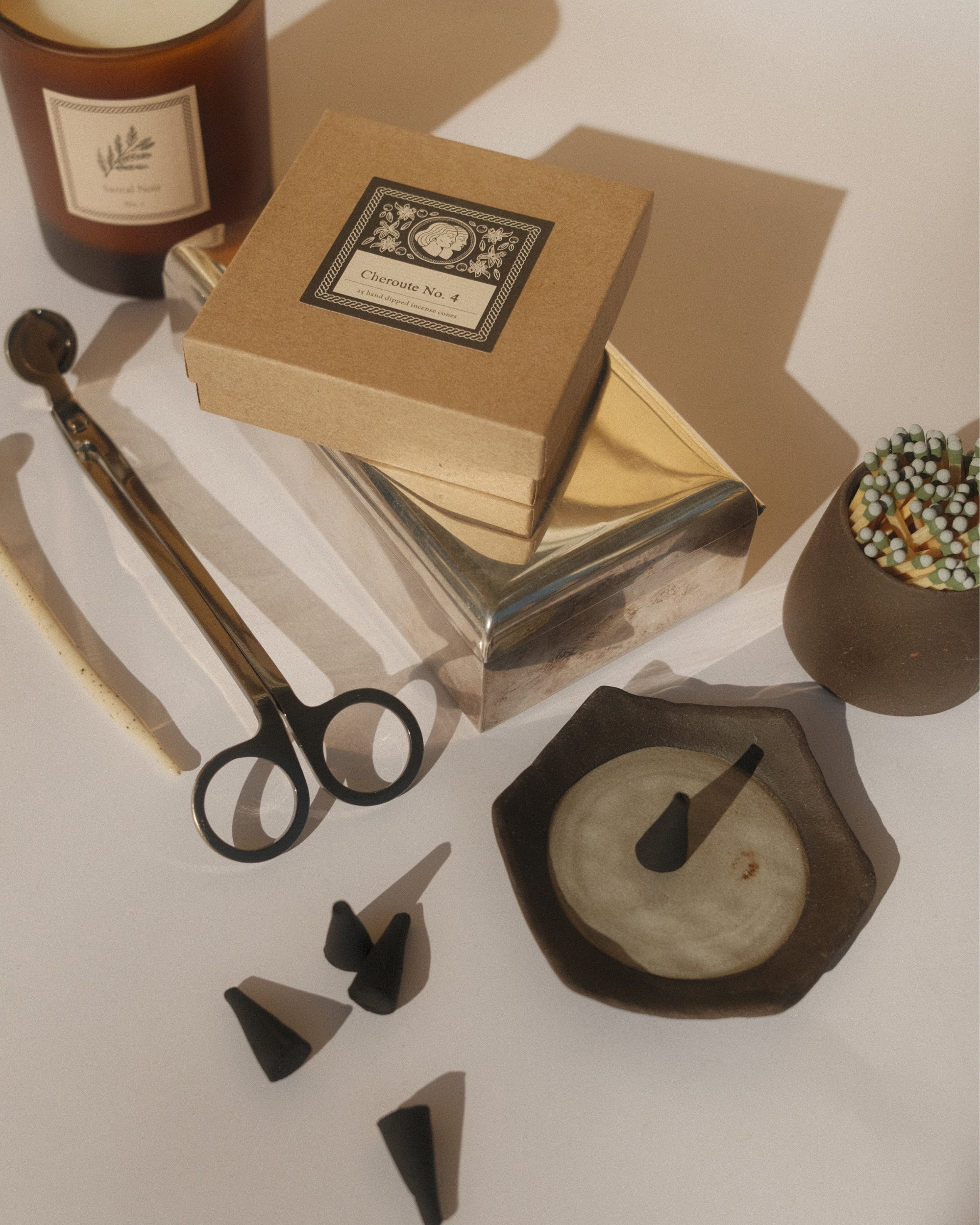 Candle tools and materials including a candle, wick trimmer, and a ceramic incense  holder on a neutral background.
