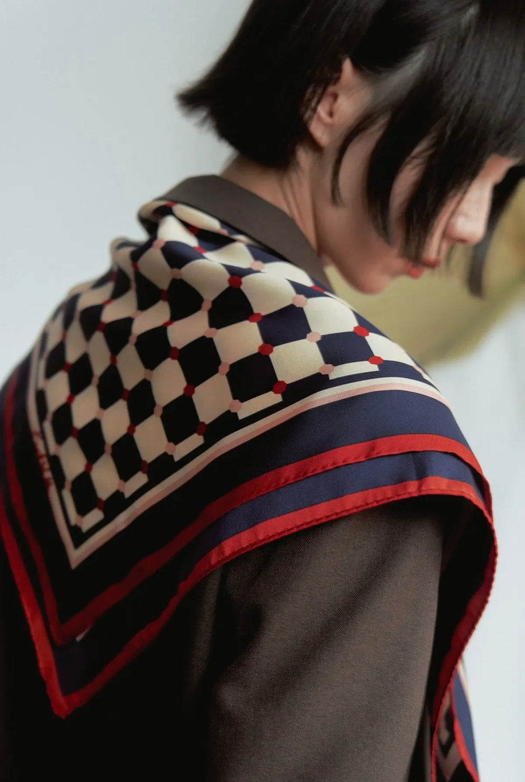 A model wearing a blue and white checkered scarf with a red border across their shoulders and a blurred background. 