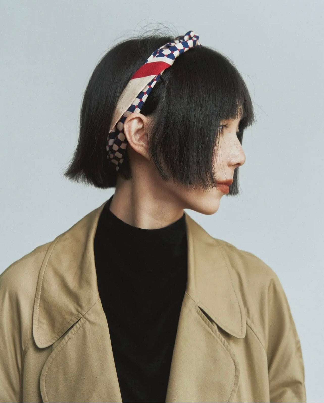 Person wearing a beige jacket and black shirt with a patterned headband on a light gray background