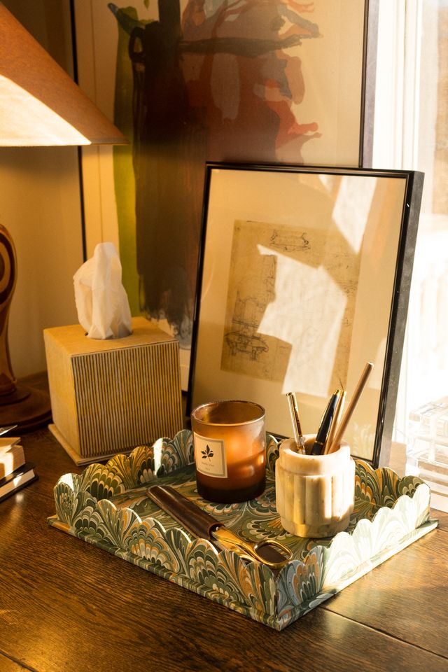 Sunlit desk setting with decorative marbled tray with a candle, a tissue box, and framed artwork on a wooden surface.
