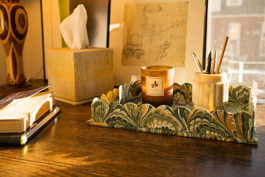 Candle on a scalloped marbled tray with stationery items on a wooden desk.