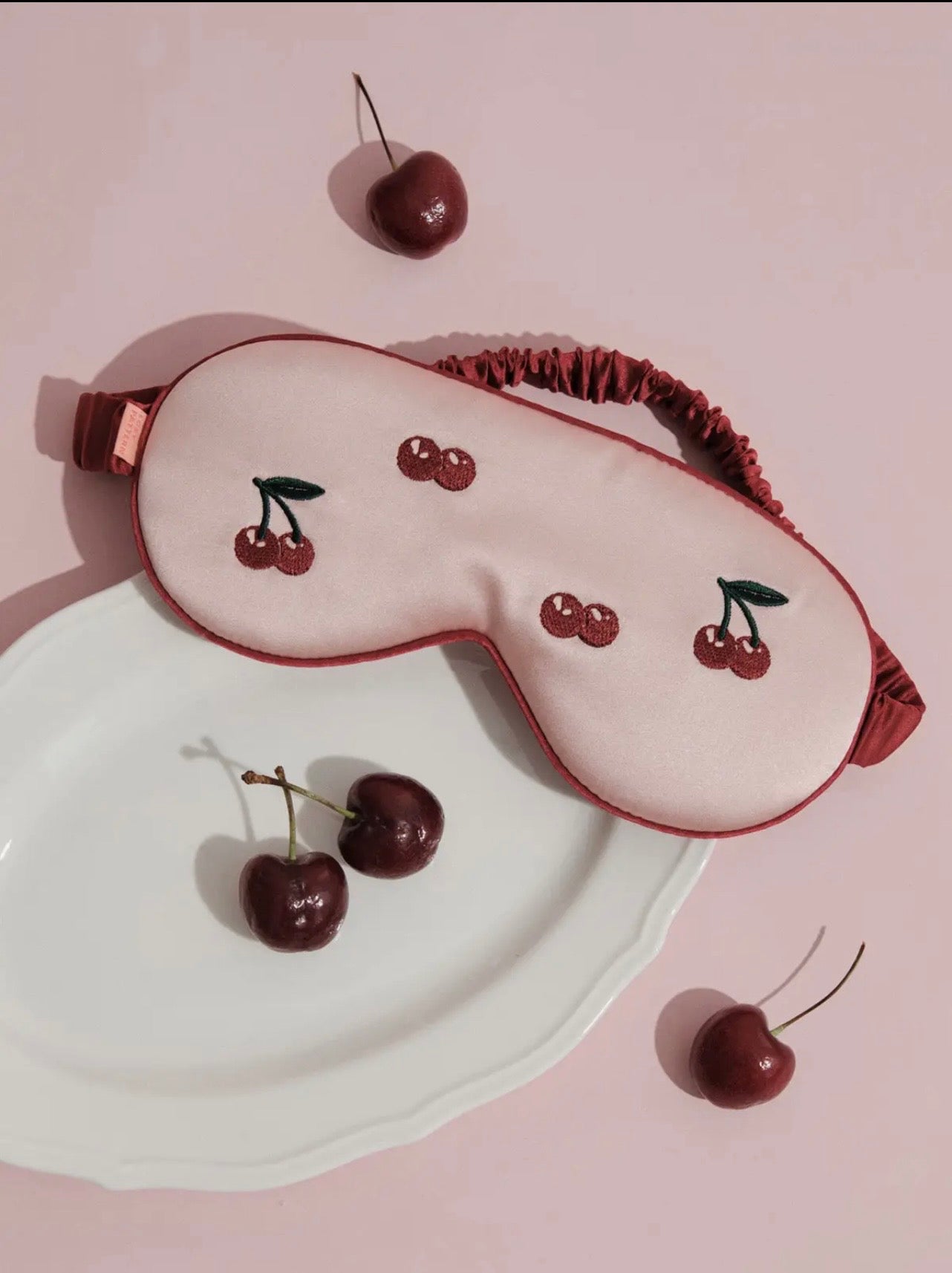 Sleep mask with cherry design on a pink background and four cherries on the side. 
