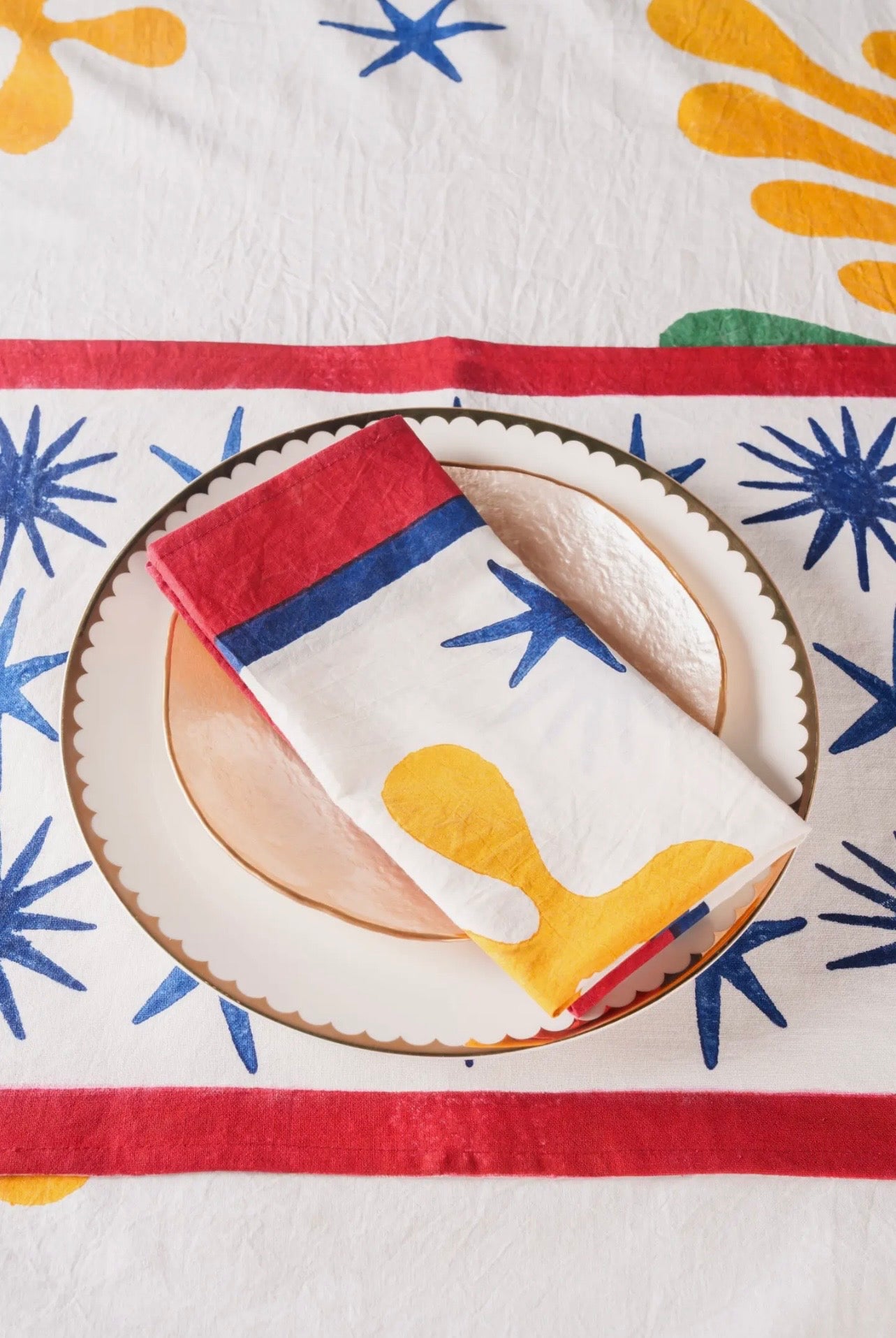 A folded cloth napkin with red top and yellow and blue pattern on a plate, on a colorful patterned tablecloth.