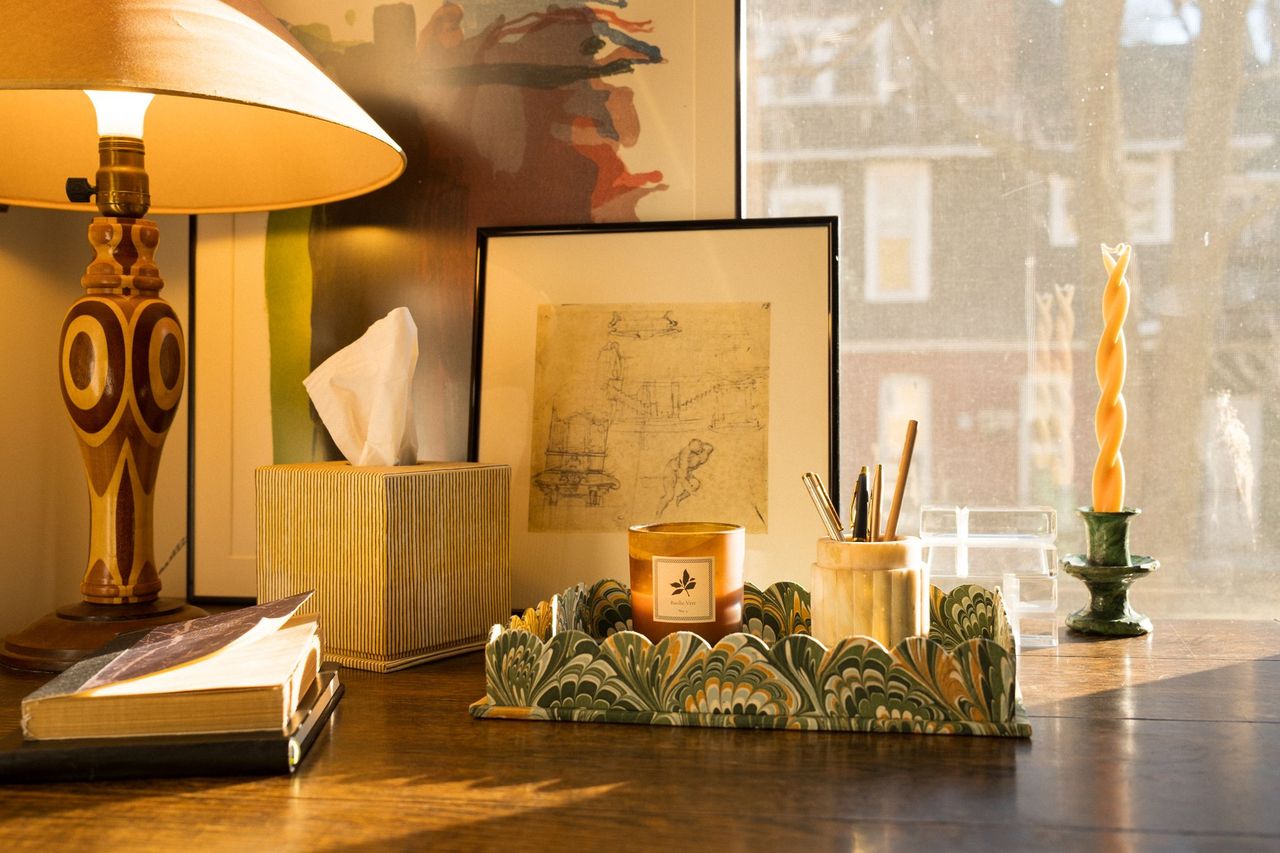 A styled sunlit desk setting with green marbled tray and striped tissue box with lamp and layered artwork. 