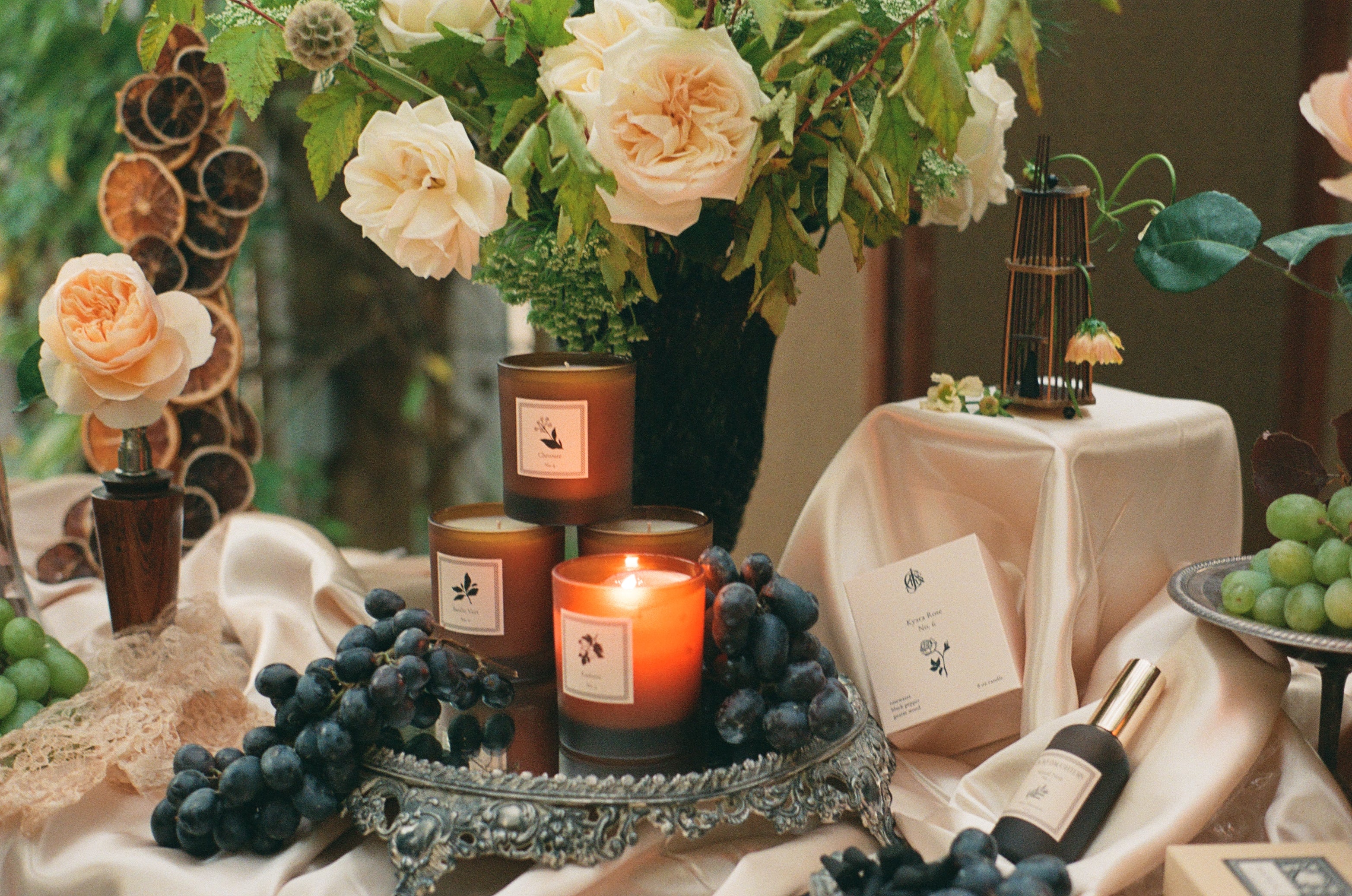 A styled tabletop scene with lit amber glass candles, a bouquet of cream roses, green grapes, dried orange slices, and a small birdcage decoration on draped satin fabric.